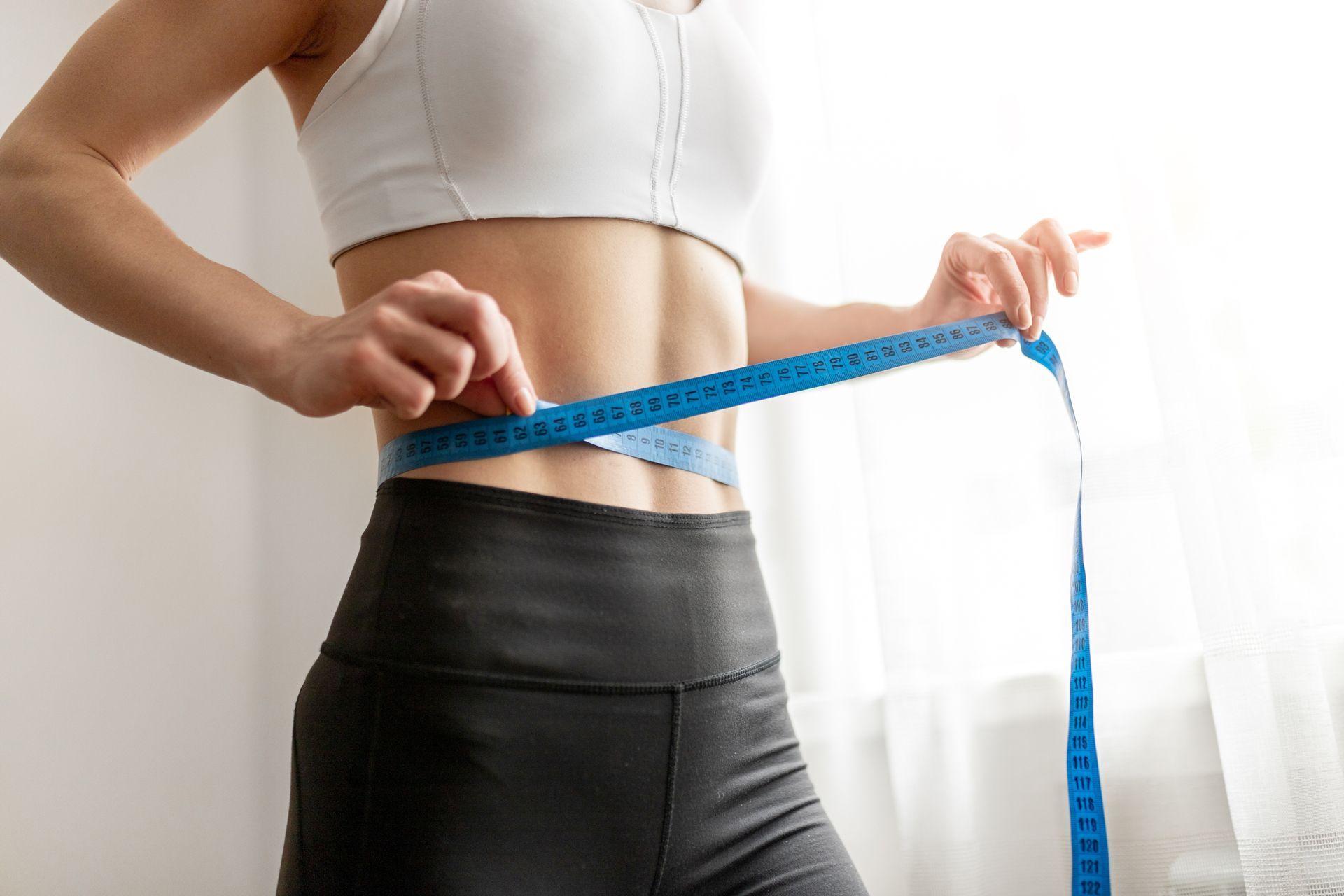 Person measuring waist with blue tape measure, wearing white top and black leggings