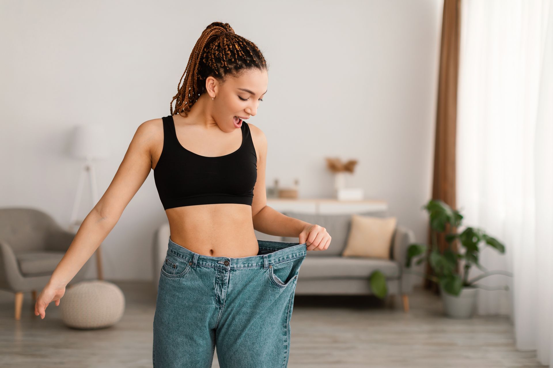 Woman standing in a living room, holding up oversized blue jeans and wearing a black crop top