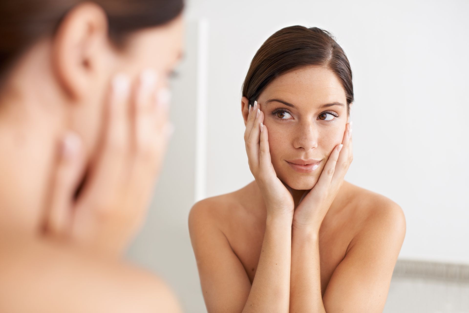 Woman gently touching her face while looking in a mirror, with her reflection visible.