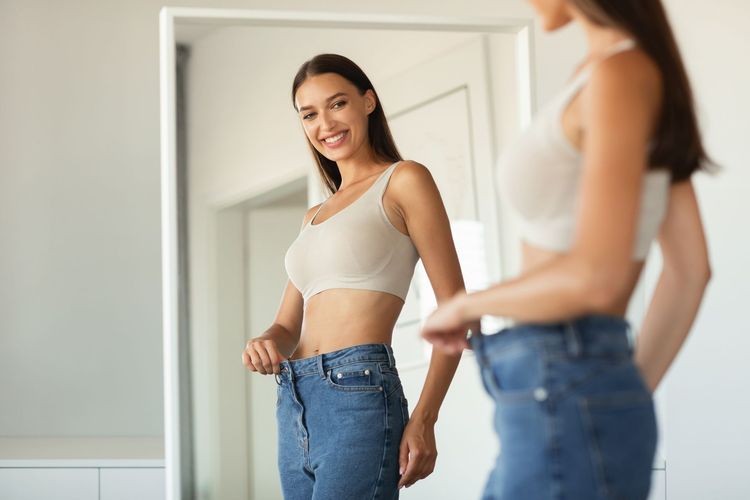 Woman smiling at a mirror while trying on jeans and a beige tank top in a bright room