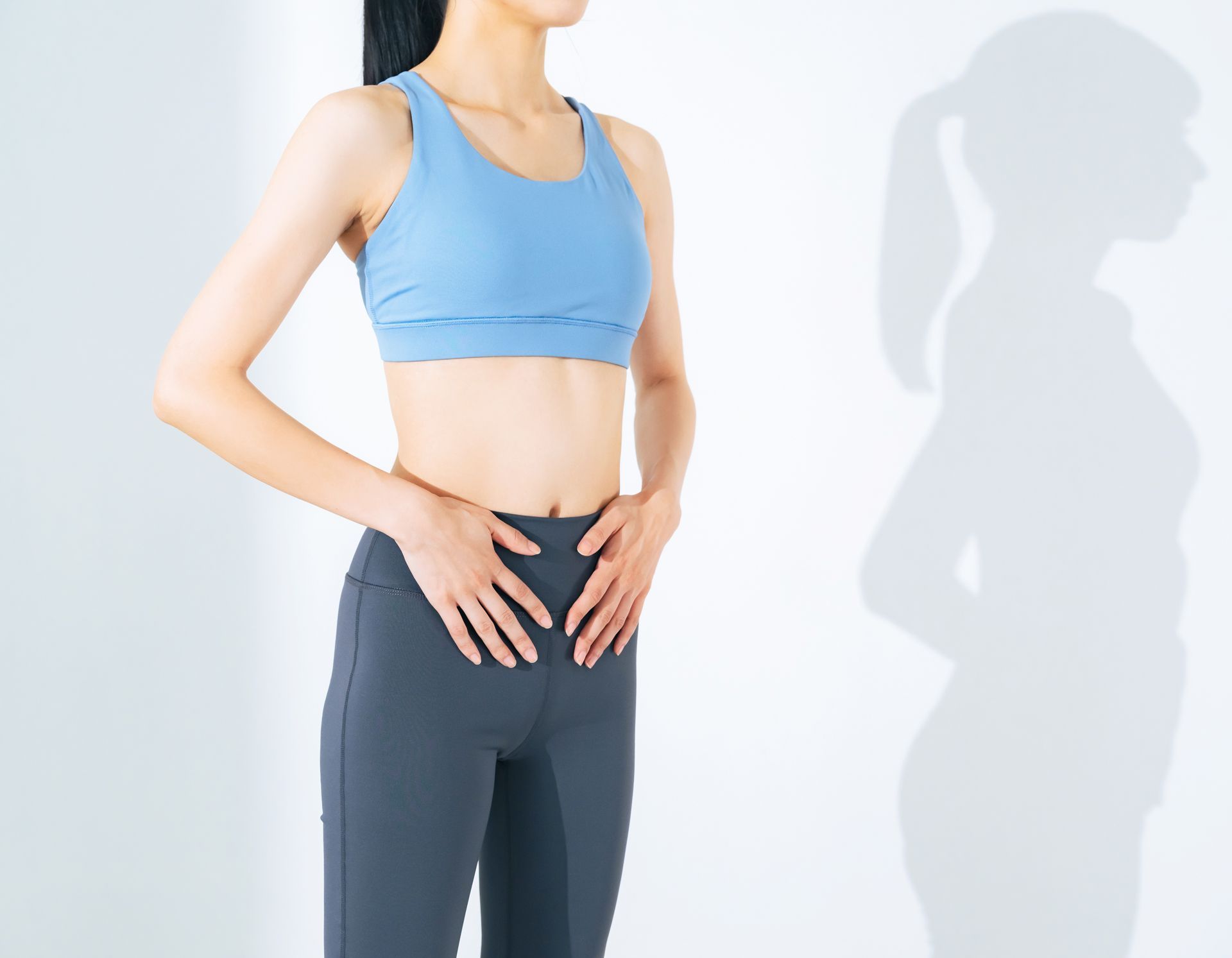 Woman in blue sports bra and gray leggings standing with hands on lower abdomen against a white wall.