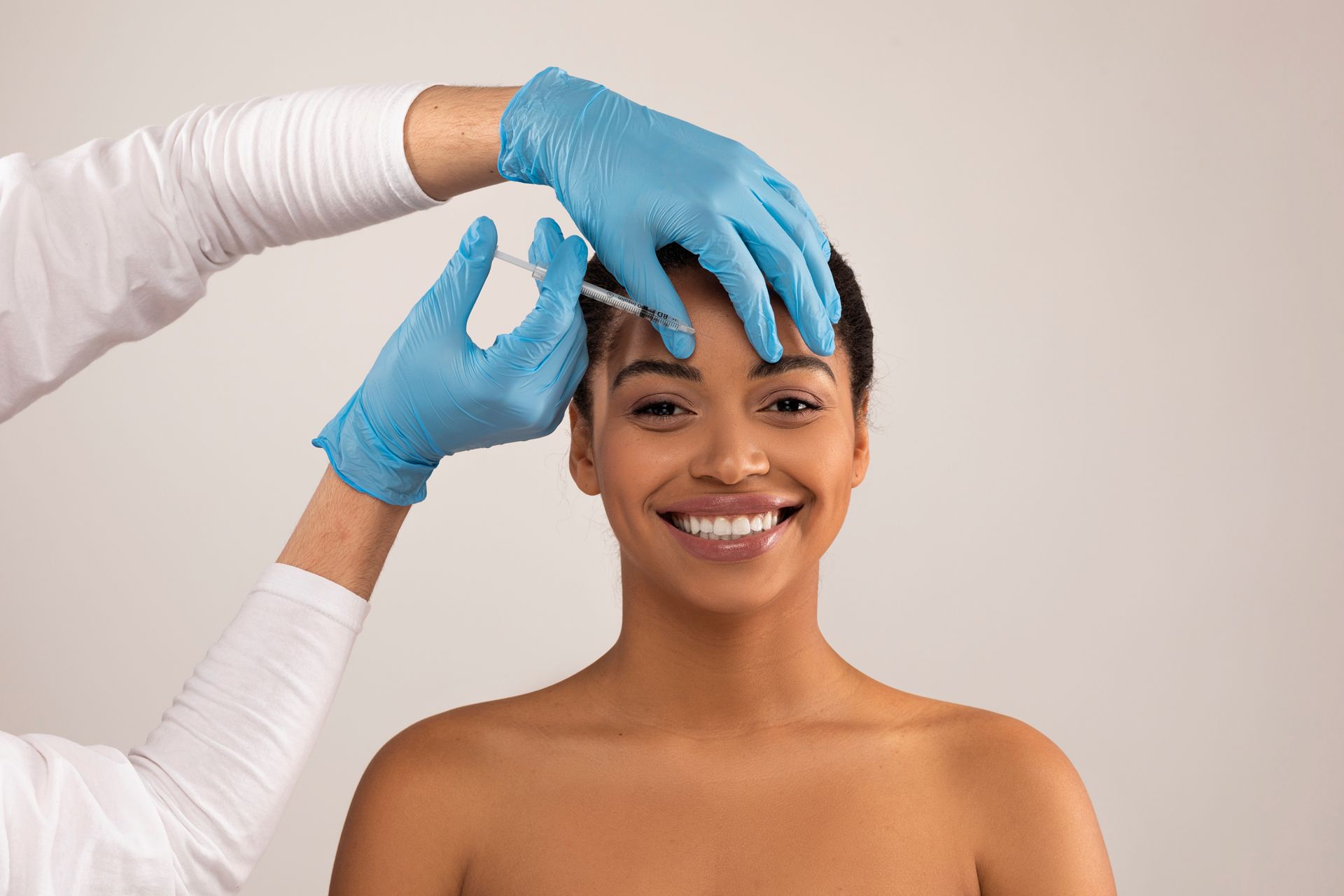 Gloved hands shape a smiling model’s eyebrows against a plain background