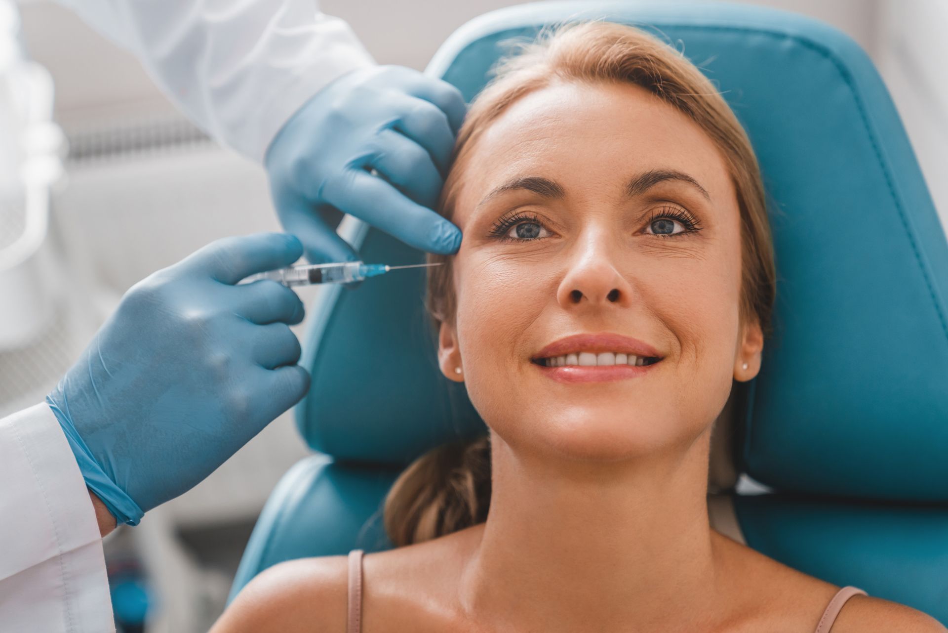 A woman smiles while receiving an injection in a medical chair, with gloved hands administering the shot.