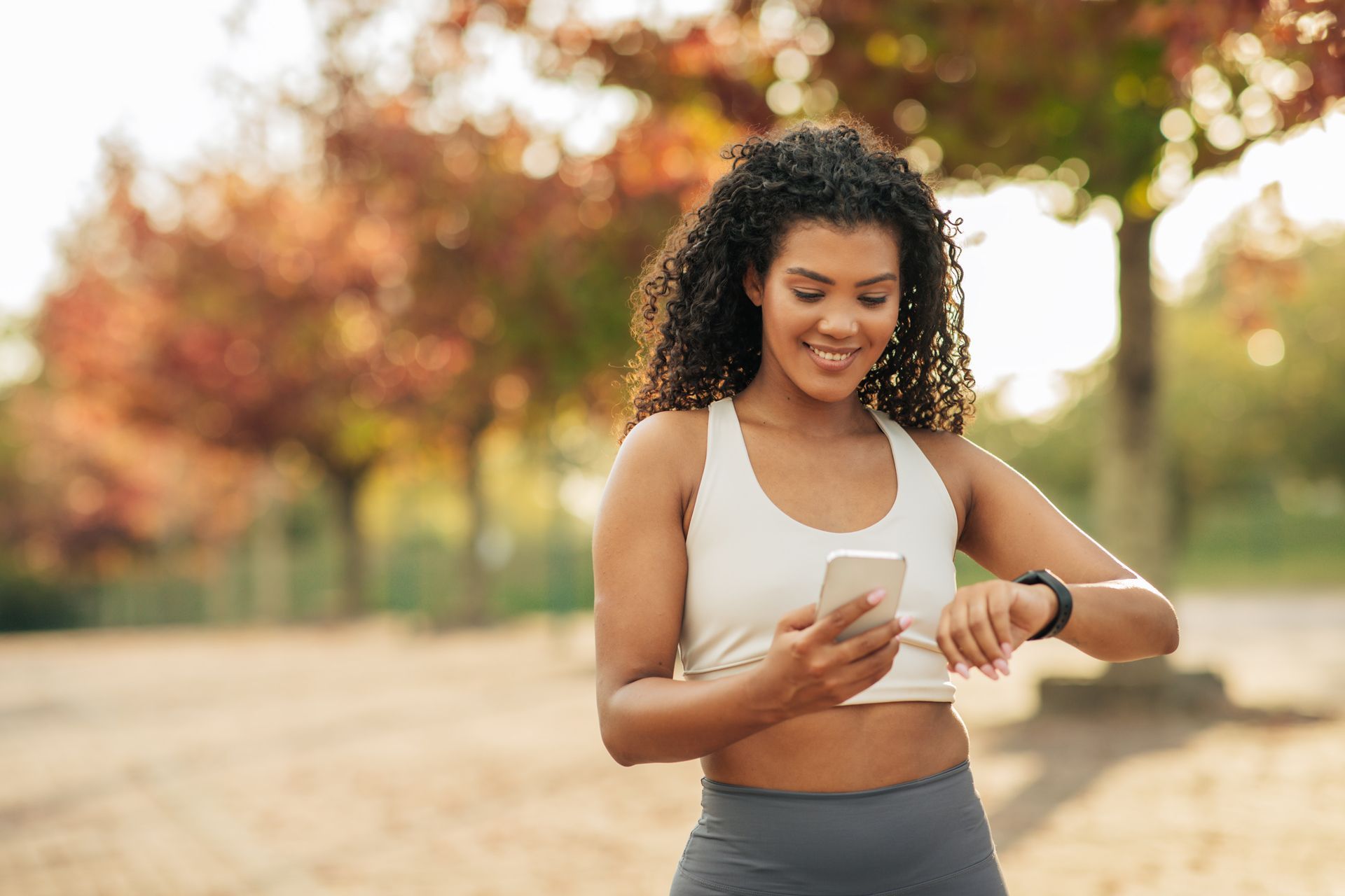 Woman checking fitness watch while holding phone outdoors in warm sunlight