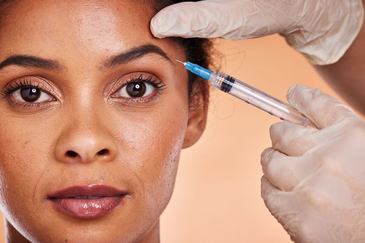 Gloved hands injecting a woman’s forehead with a syringe against a peach background