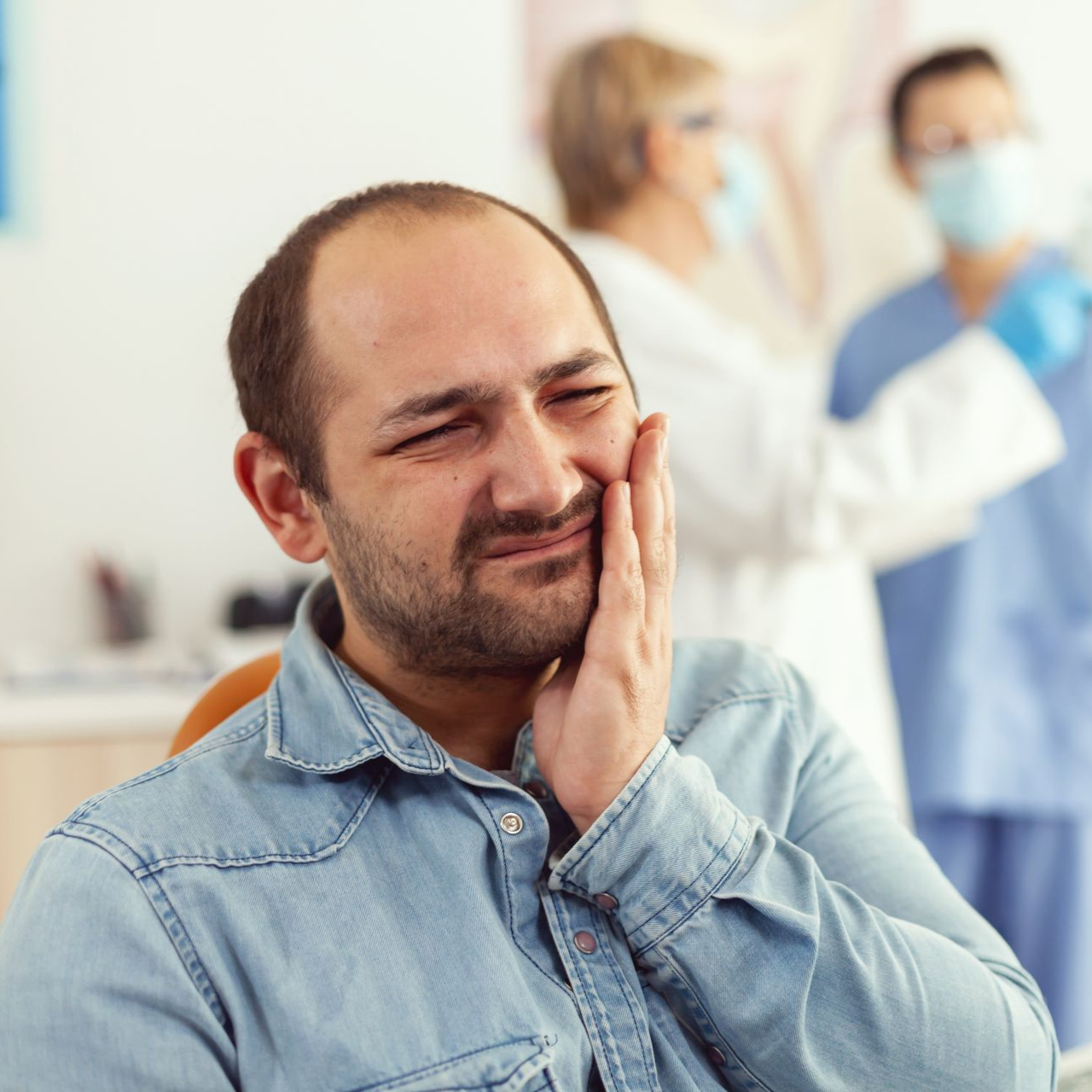 Man holding his cheek in a clinic waiting room, looking uncomfortable; medical staff blurred in background