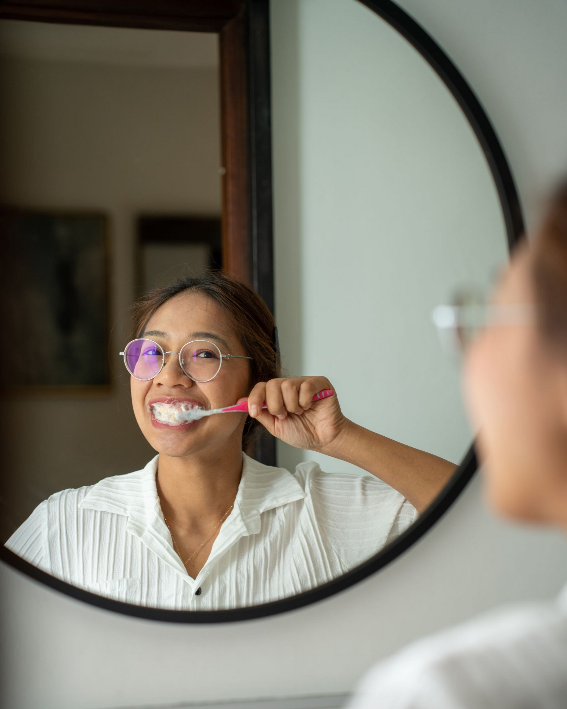 Woman brushing her teeth in a bathroom mirror, wearing glasses and a white shirt.