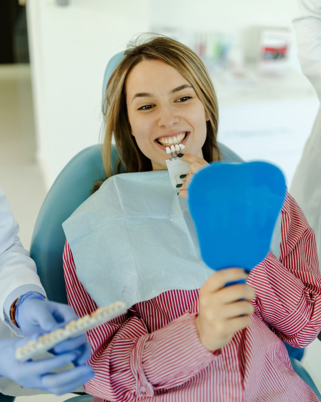 Woman smiling during a dental exam, holding a blue mirror while a dentist works nearby.