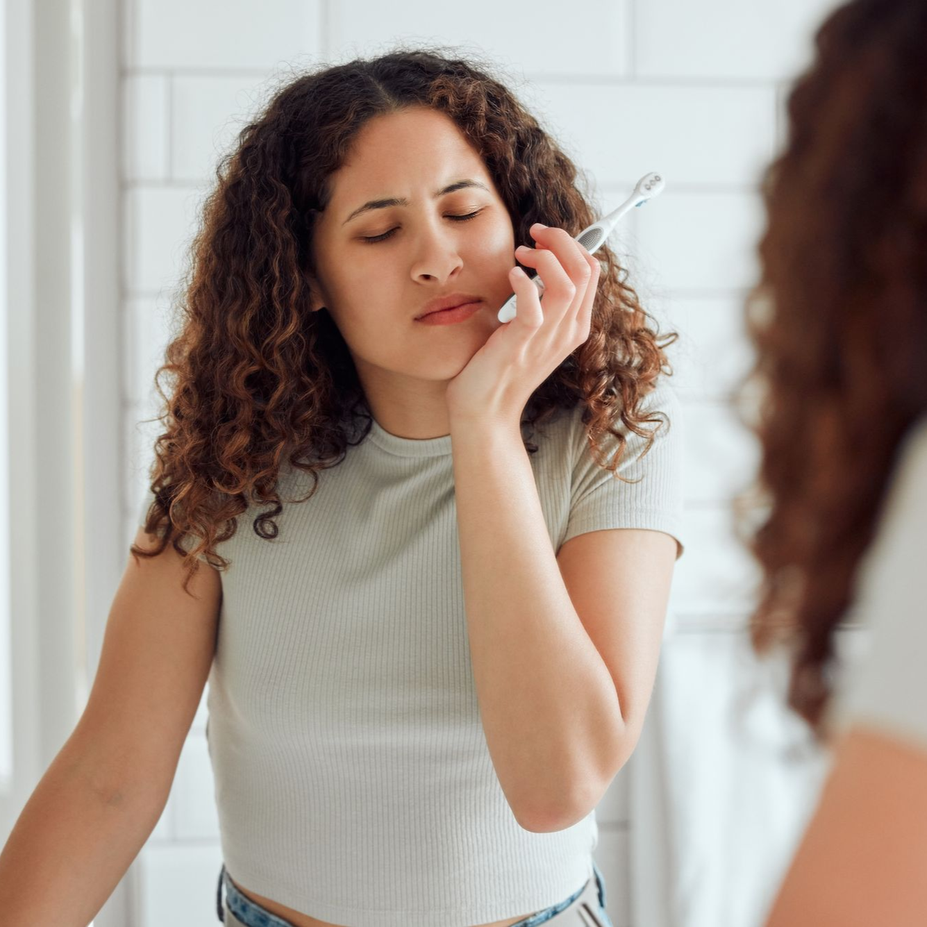 Woman with curly hair touching her cheek while looking in a bathroom mirror