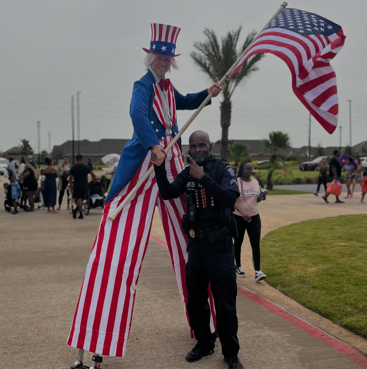 Person on stilts dressed as Uncle Sam with officer pointing, holding flag. Outdoor event with people.
