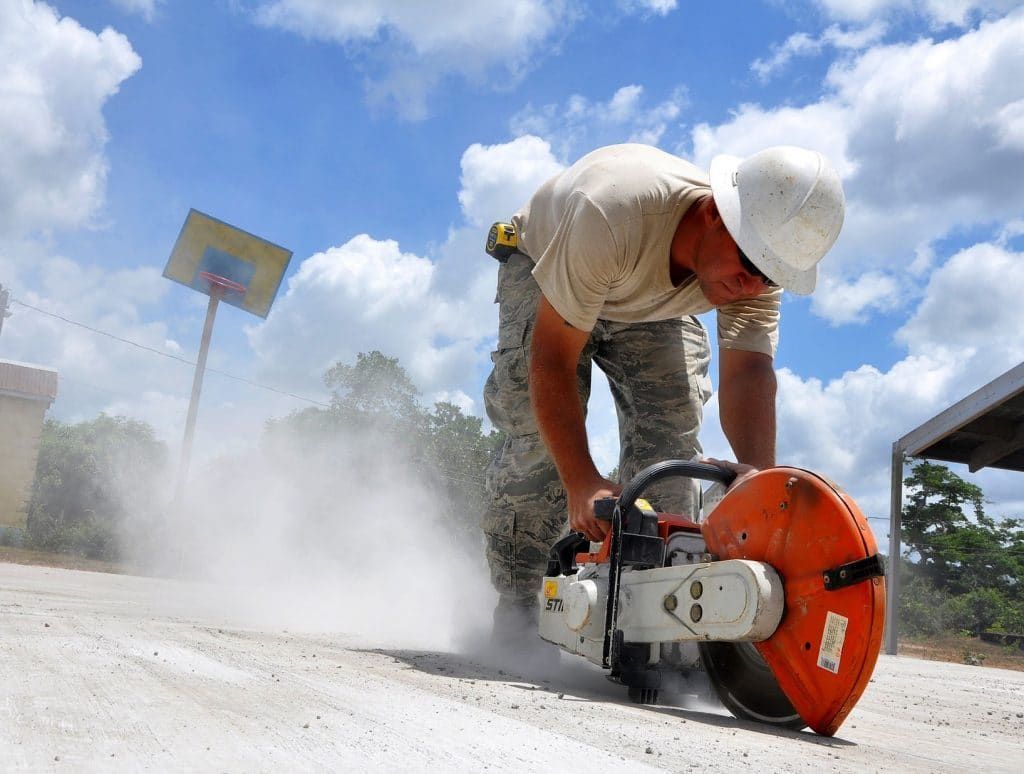A worker in a hard hat uses a large orange power saw to cut into a concrete surface, creating a cloud of dust outdoors.