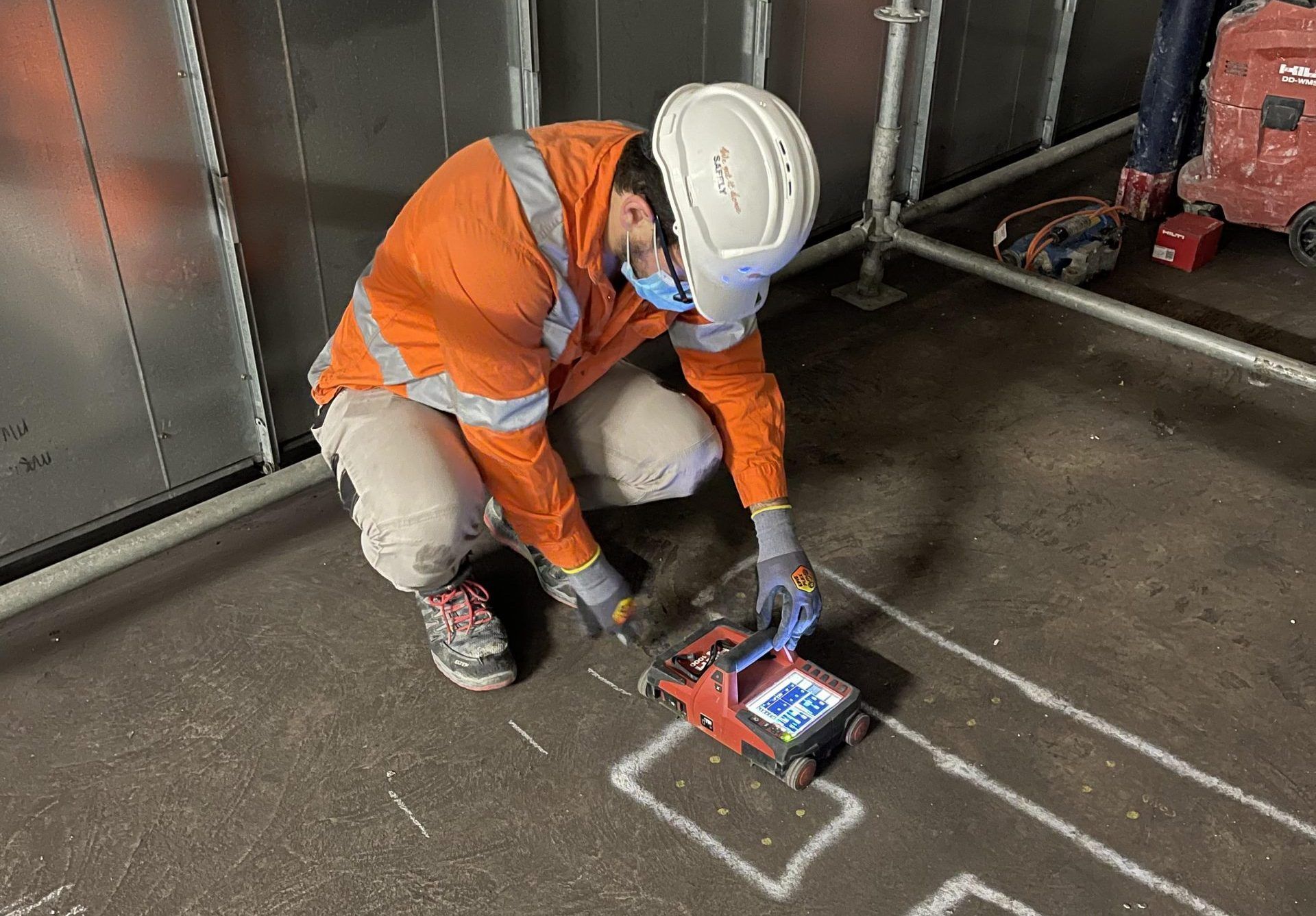 Construction worker in orange vest and hard hat using a red scanning device on a concrete floor.