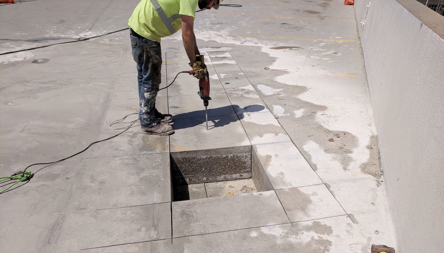 Worker using a jackhammer beside an open utility trench on a sidewalk.