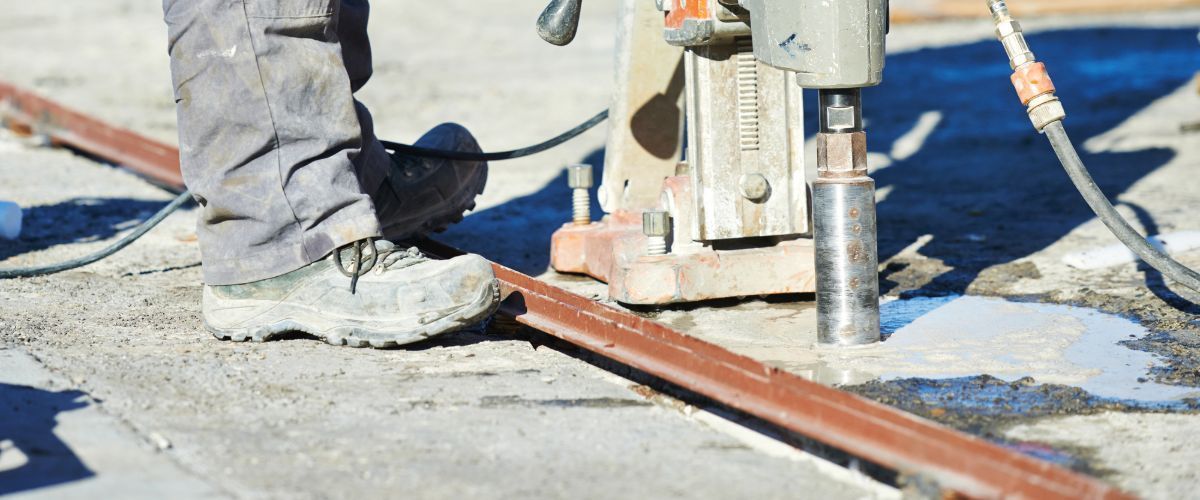 A worker operates a diamond core drill on a concrete surface next to a metal rail, with water pooling at the drill site.