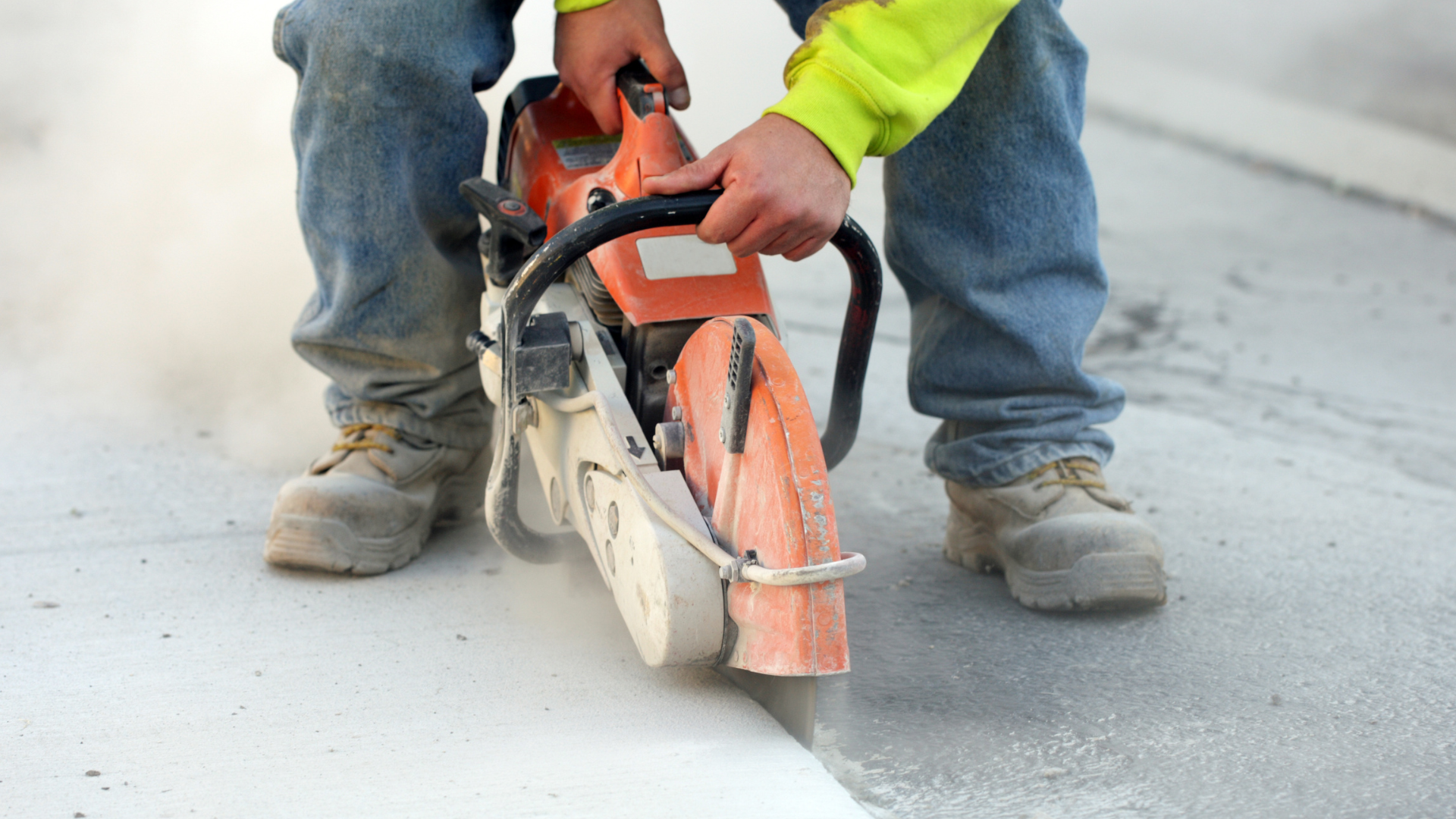 Person cutting concrete with a power saw, creating dust, wearing work boots and jeans.