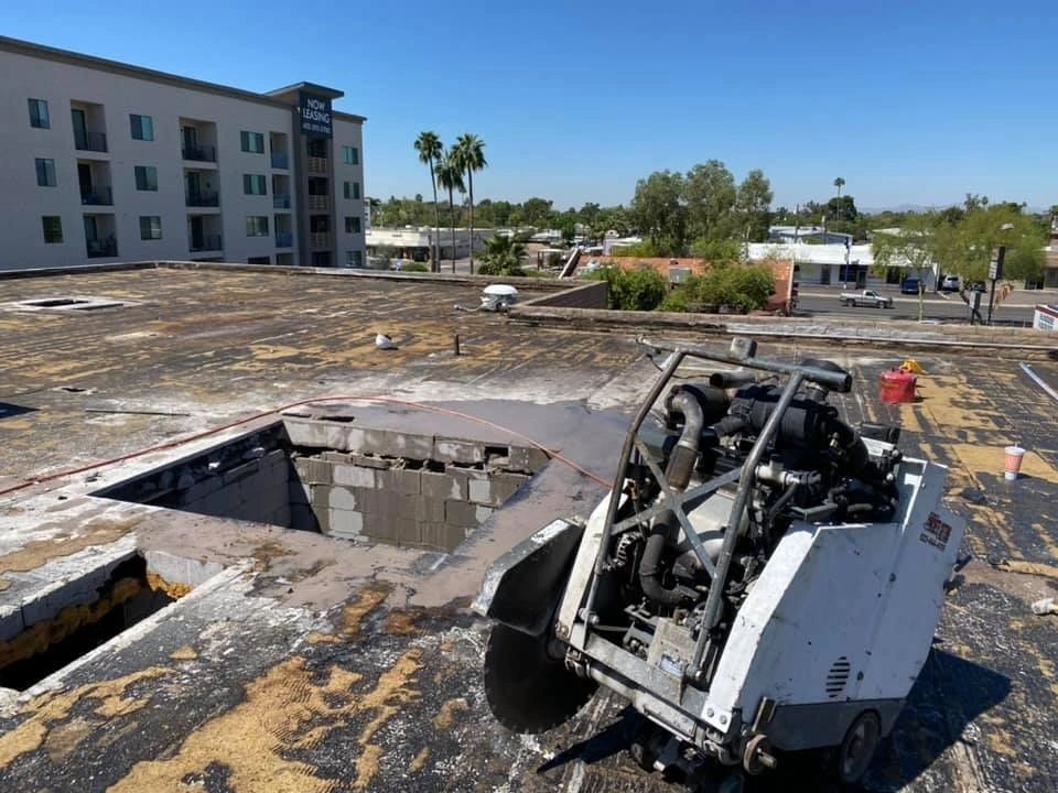 Roof demolition: Saw cutting through asphalt. Large machine on a flat roof, near a hole with a building in background.