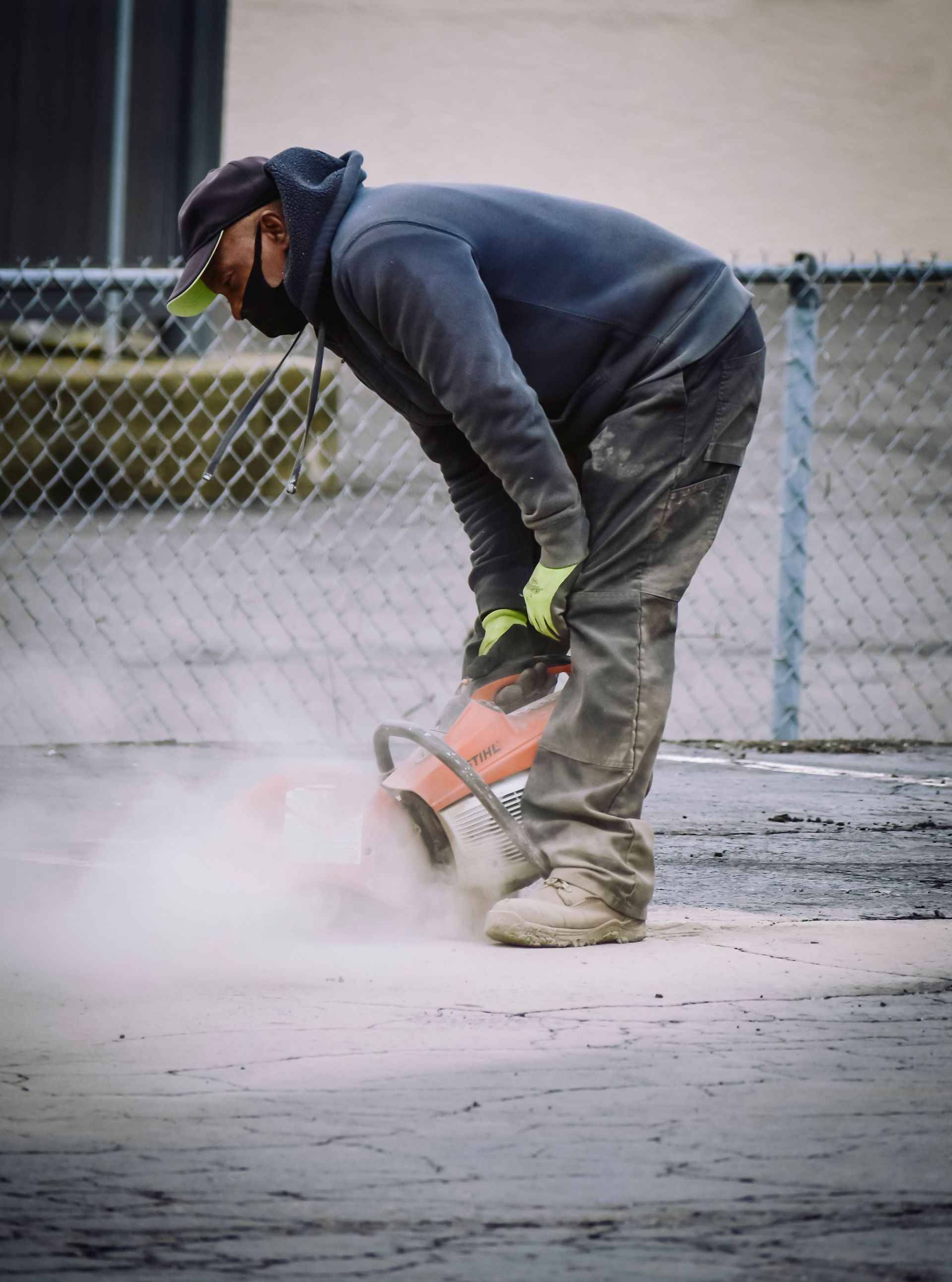Worker cutting concrete with a handheld saw outdoors, wearing dark hoodie and gloves.