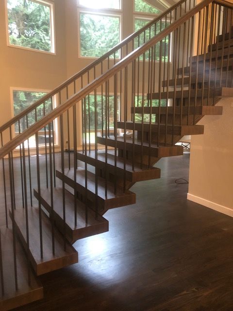 Floating wooden staircase with dark metal railings, light wooden handrail, and windows in the background.