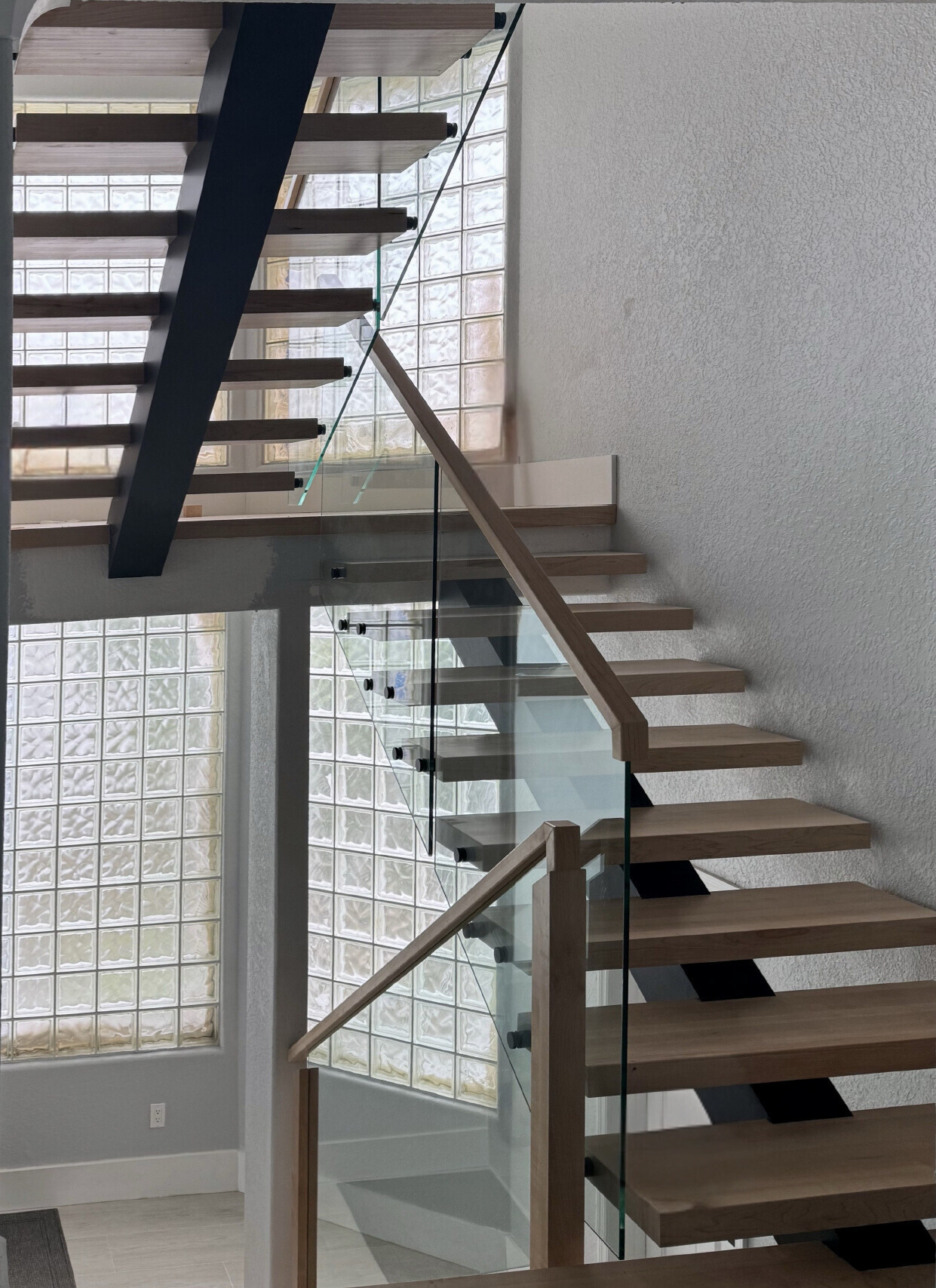 Wooden floating staircase with glass railing against a textured wall and glass block window.
