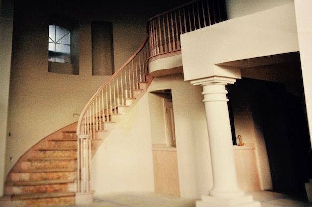 Curved wooden staircase with banister, ascending against a beige wall, a classical column supporting a landing.