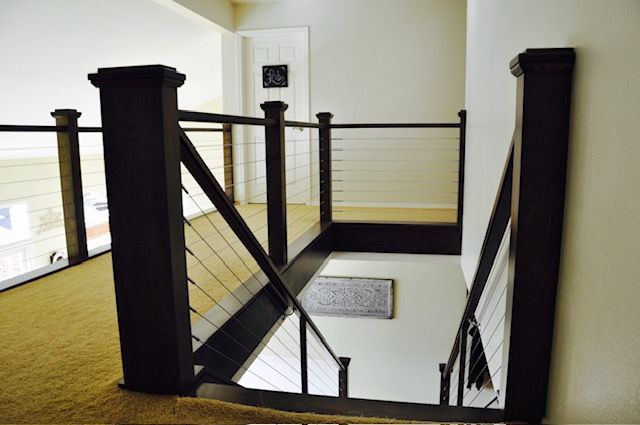 Interior view of a staircase with dark wooden posts and cable railings. Beige carpet and white walls.