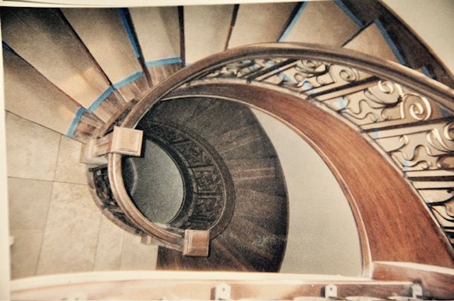 Spiral wooden staircase viewed from above. Brown wood handrail with carved details and tan steps.