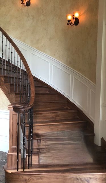 Wooden curved staircase with decorative iron balusters, white paneling, and wall sconces.