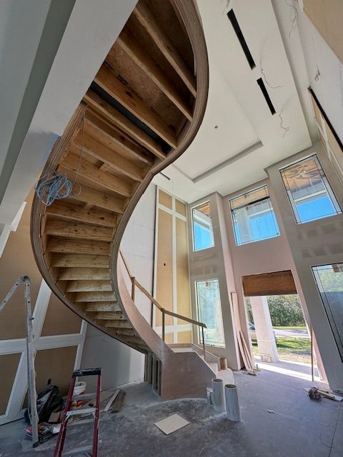 Curving wooden staircase under construction in a spacious interior, with large windows and unfinished walls.