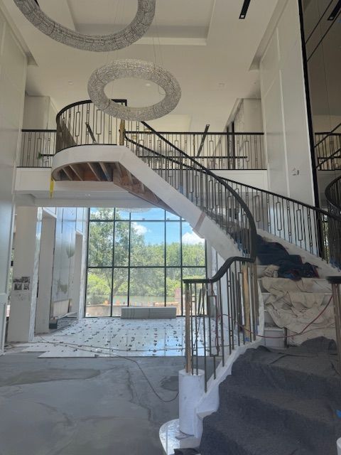 Grand, curved staircase in a building under construction, with high ceilings, large windows, and a circular chandelier.