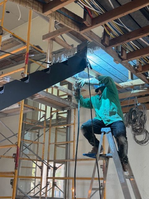 Person welding metal overhead while standing on a ladder inside a building under construction.