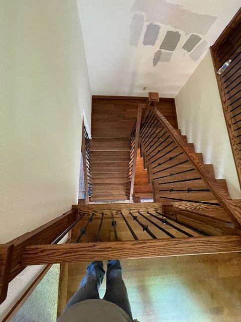View from top of wooden staircase with dark railing, light walls. Person's feet visible at the bottom.