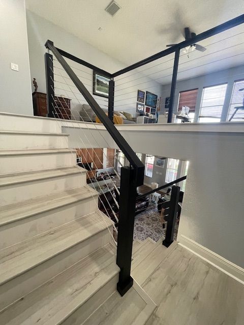 Staircase with black railing and wire cables. Gray wood-look steps lead to a landing overlooking a living area.