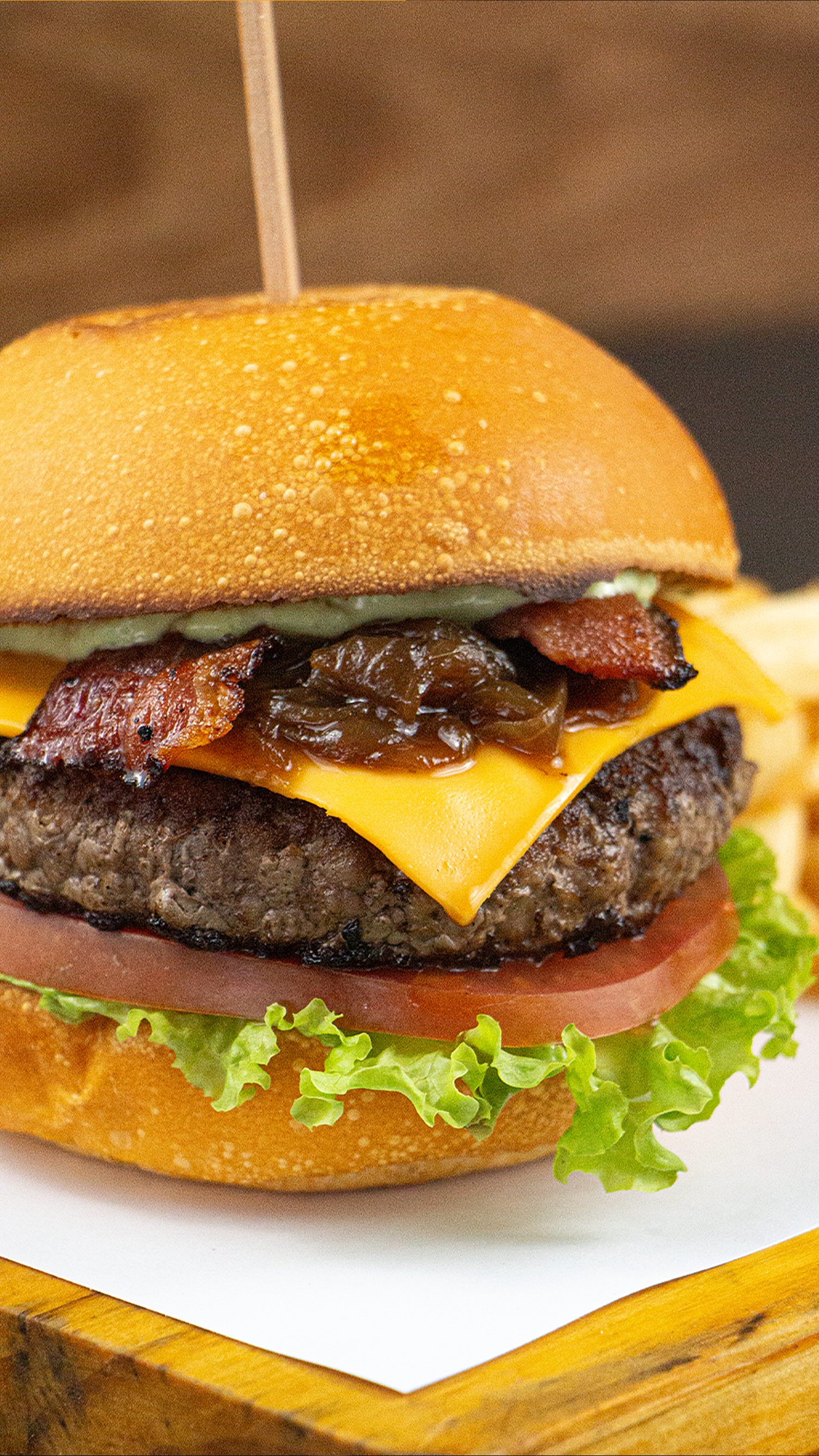 A close up of a hamburger with cheese , bacon , lettuce and tomato on a wooden table.