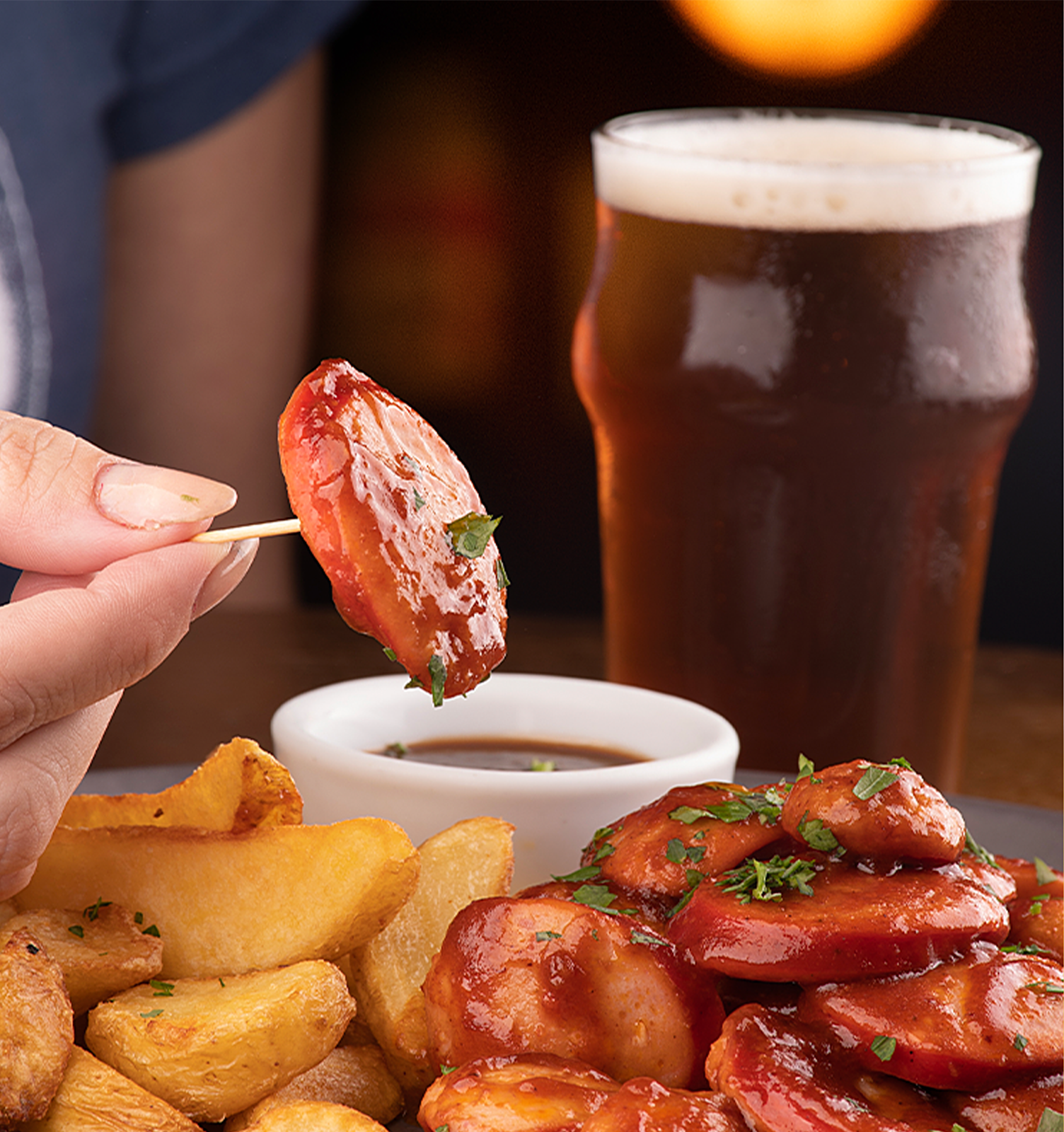 Travessa A person is dipping a sausage into a dipping sauce next to a glass of beer