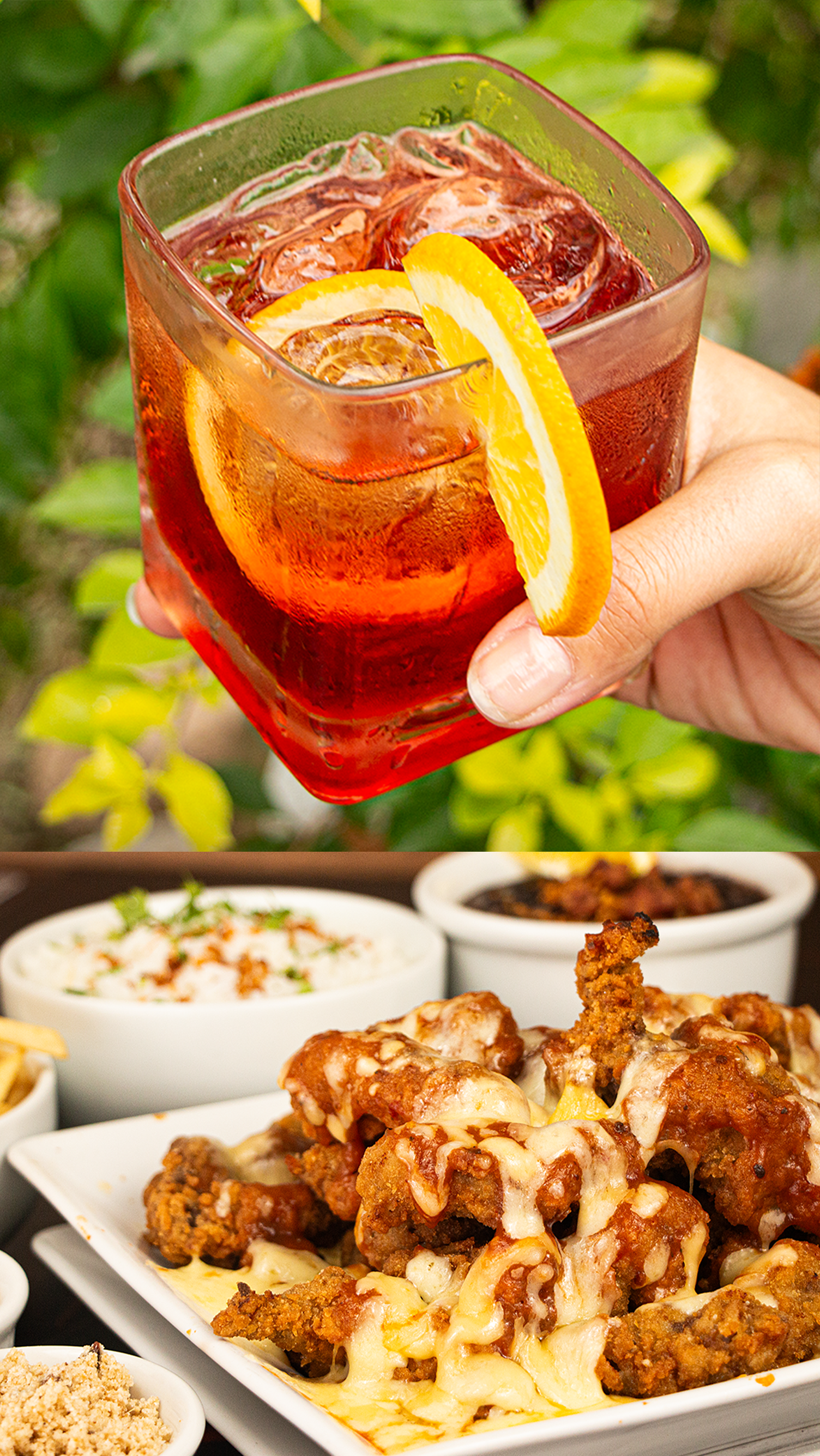 A person is holding a glass of red liquid with an orange slice in front of a plate of food.