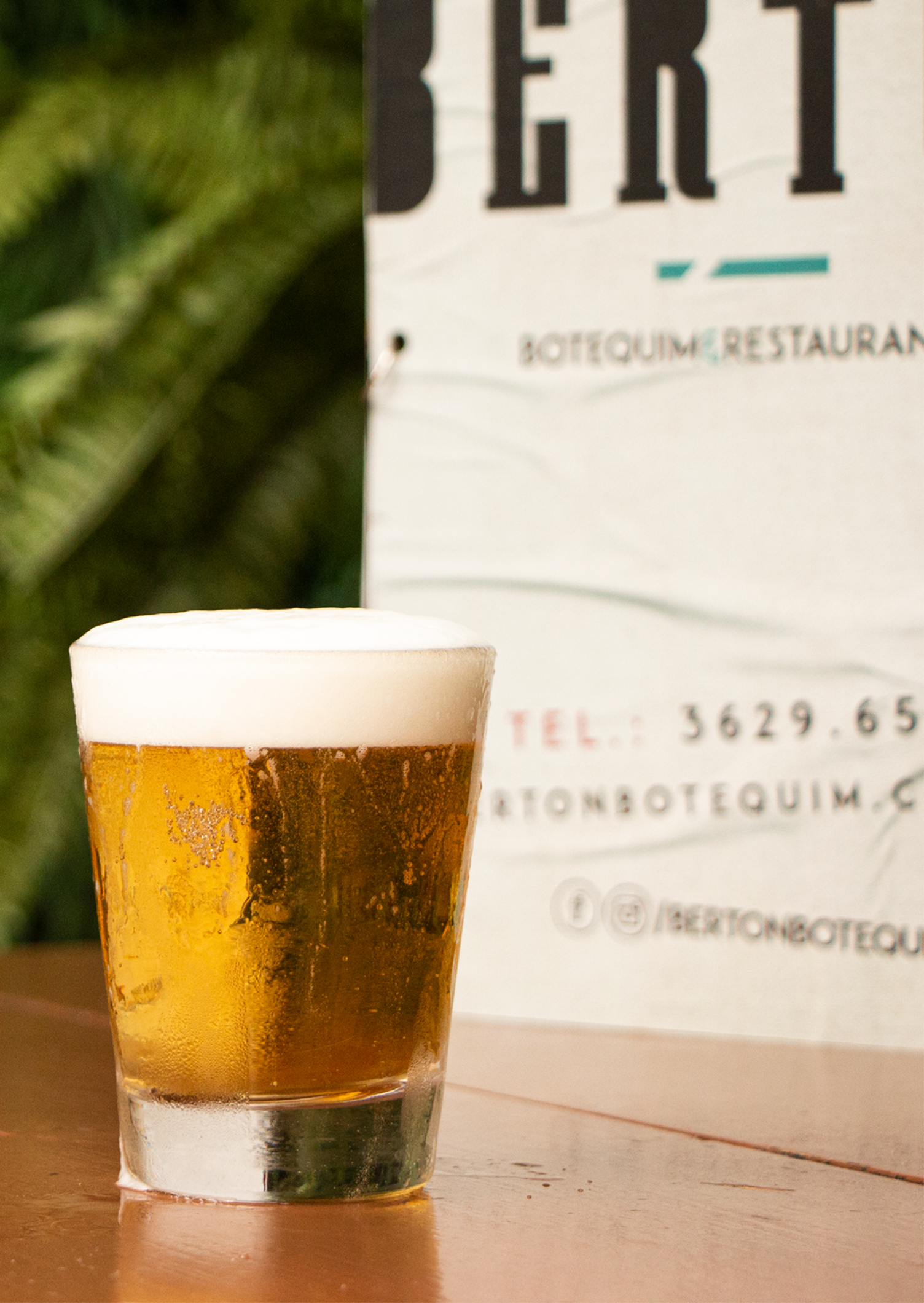Berton Botequim e Restaurante Um copo de cerveja está sobre uma mesa em frente a uma placa que diz bert