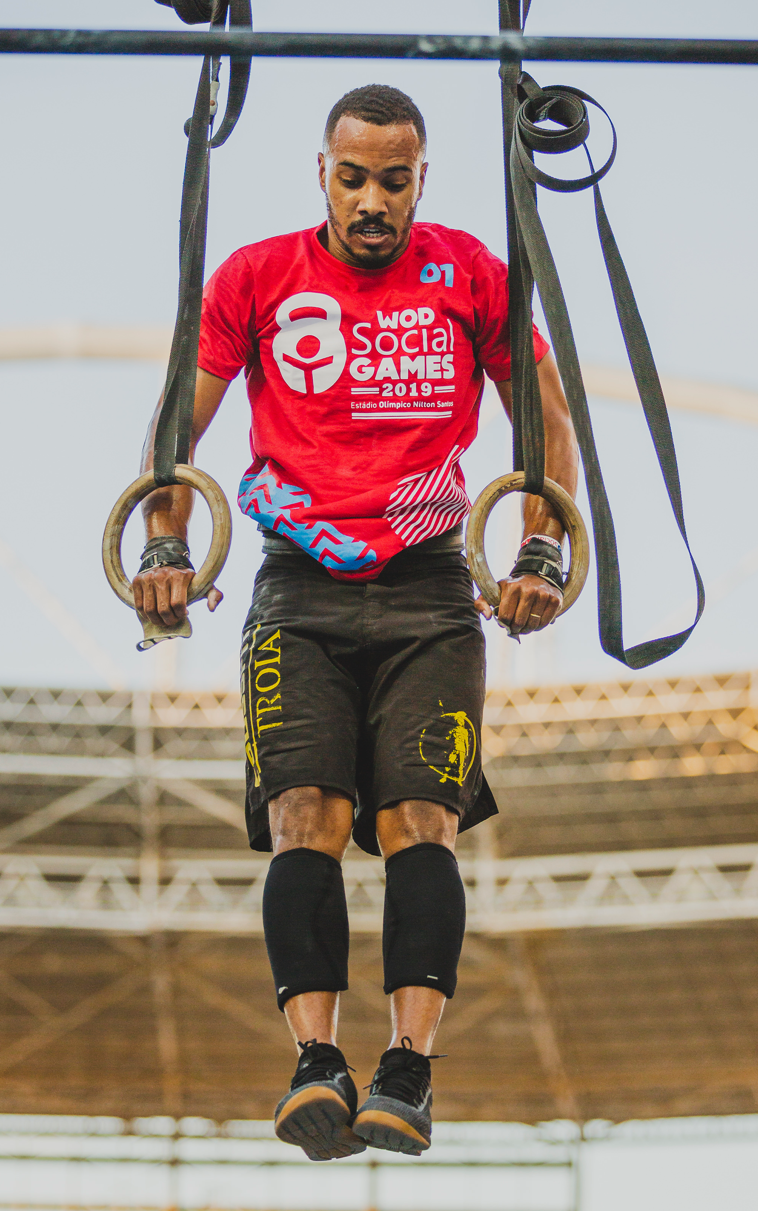 Wod Social Games Crossfit Um homem de camisa vermelha está pendurado em argolas de ginástica.