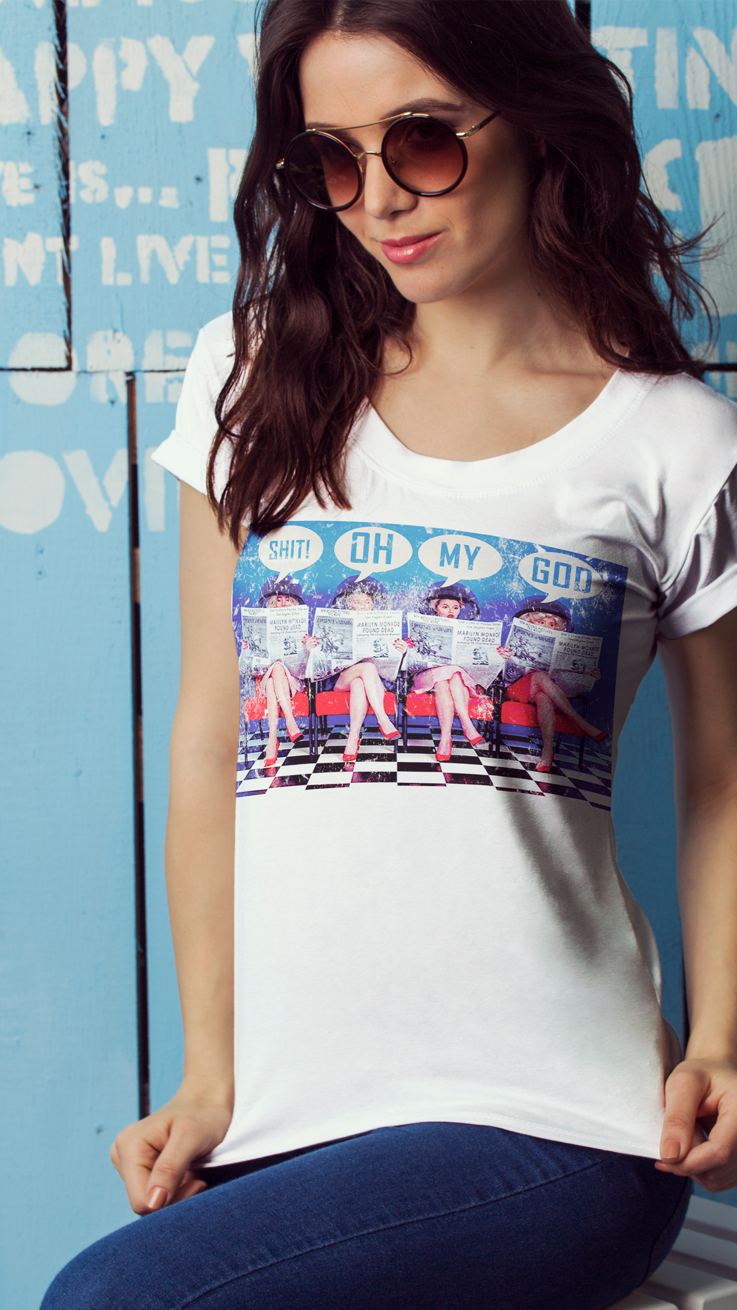 Uma mulher usando óculos escuros e uma camiseta branca está sentada em frente a uma parede azul.
