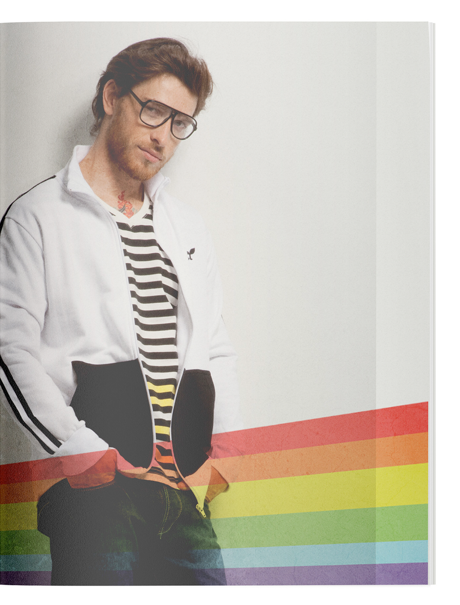 A man wearing glasses and a striped shirt is standing in front of a rainbow flag