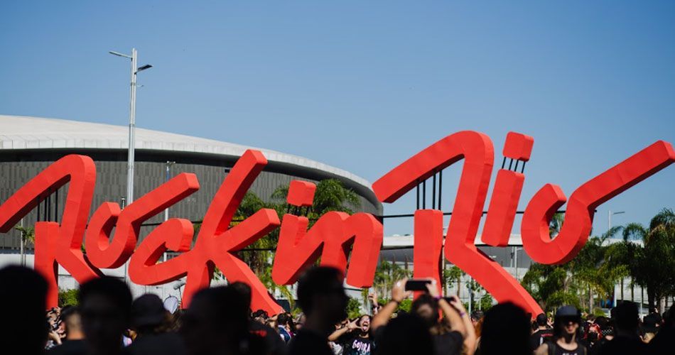 Uma multidão de pessoas está em frente a uma grande placa vermelha que diz rock in rio.