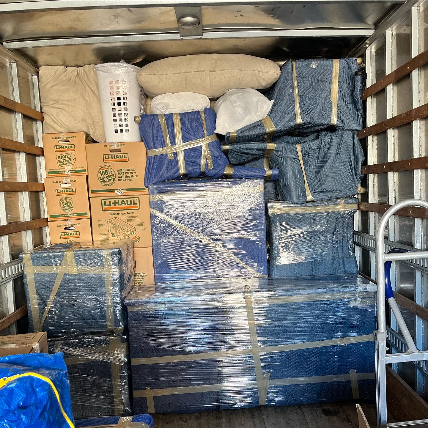 Inside a moving truck packed with furniture wrapped in blue plastic, boxes, pillows and bags.