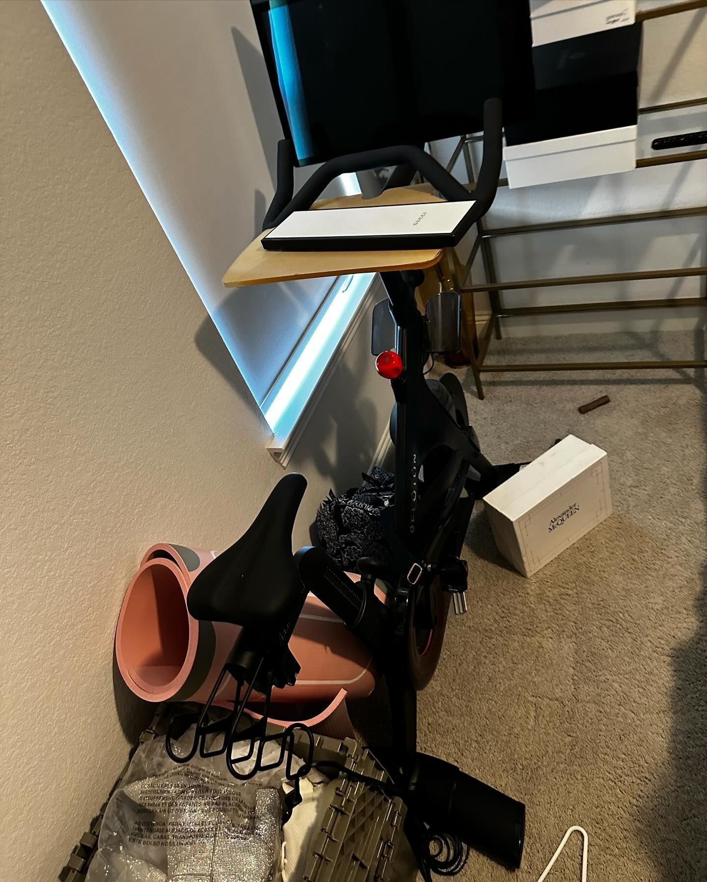 Stationary exercise bike with screen and attached shelf in a room with a white box and pink bucket.