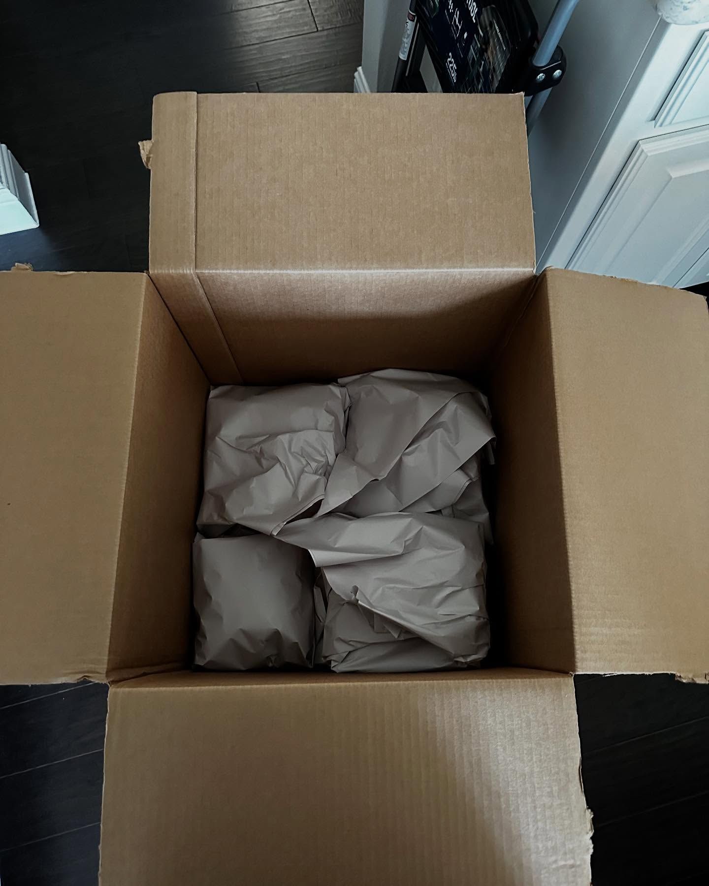 Open cardboard box with brown paper-wrapped items inside, on a dark floor.