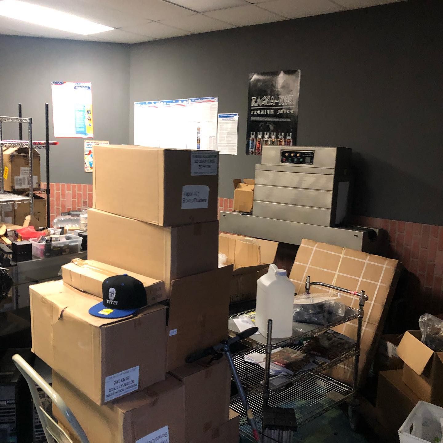 Boxes piled in a cluttered storage room with a cash register, shelving, and posters on the wall.