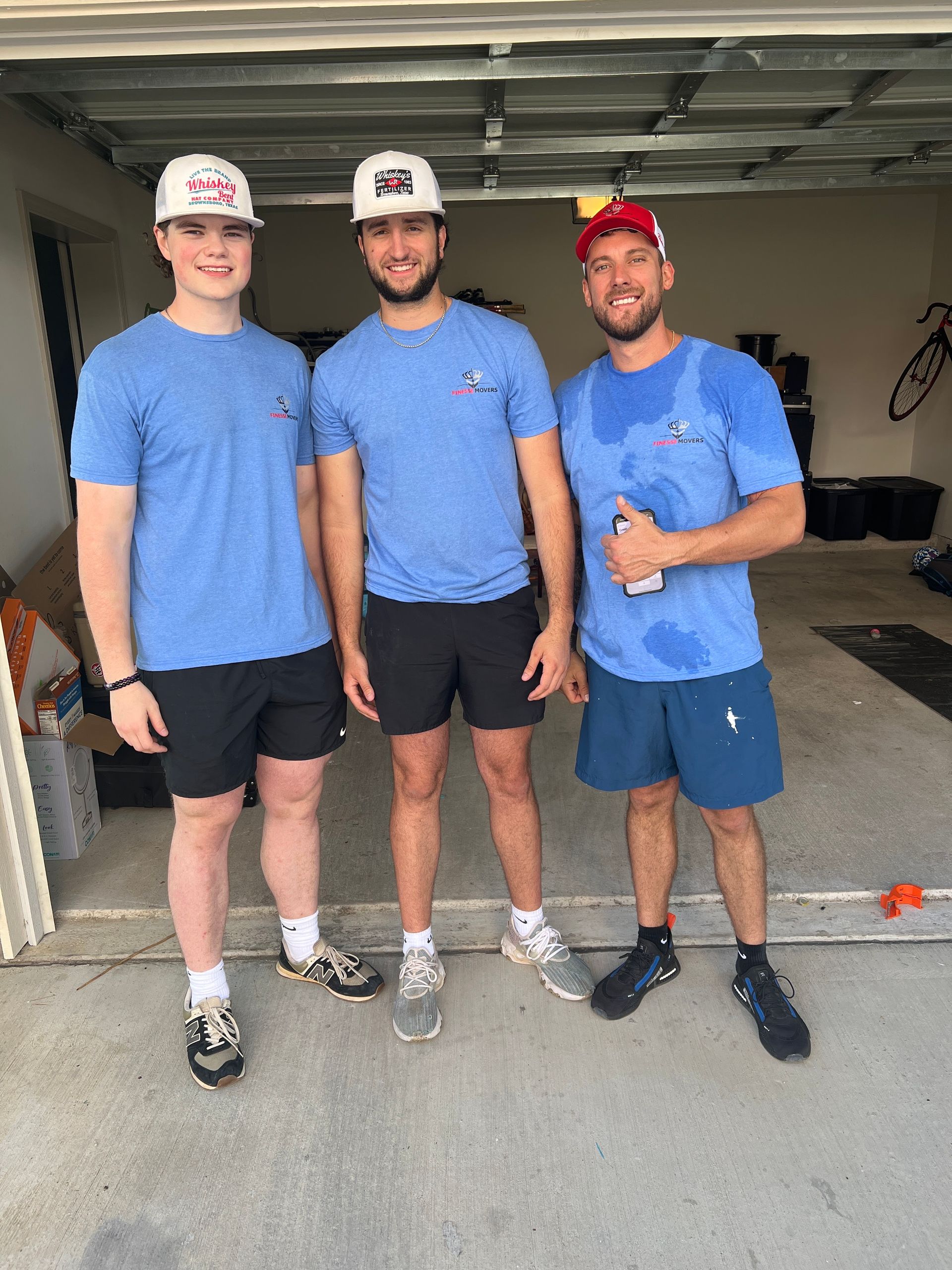 Three men wearing blue shirts, shorts, and hard hats, standing in a garage.