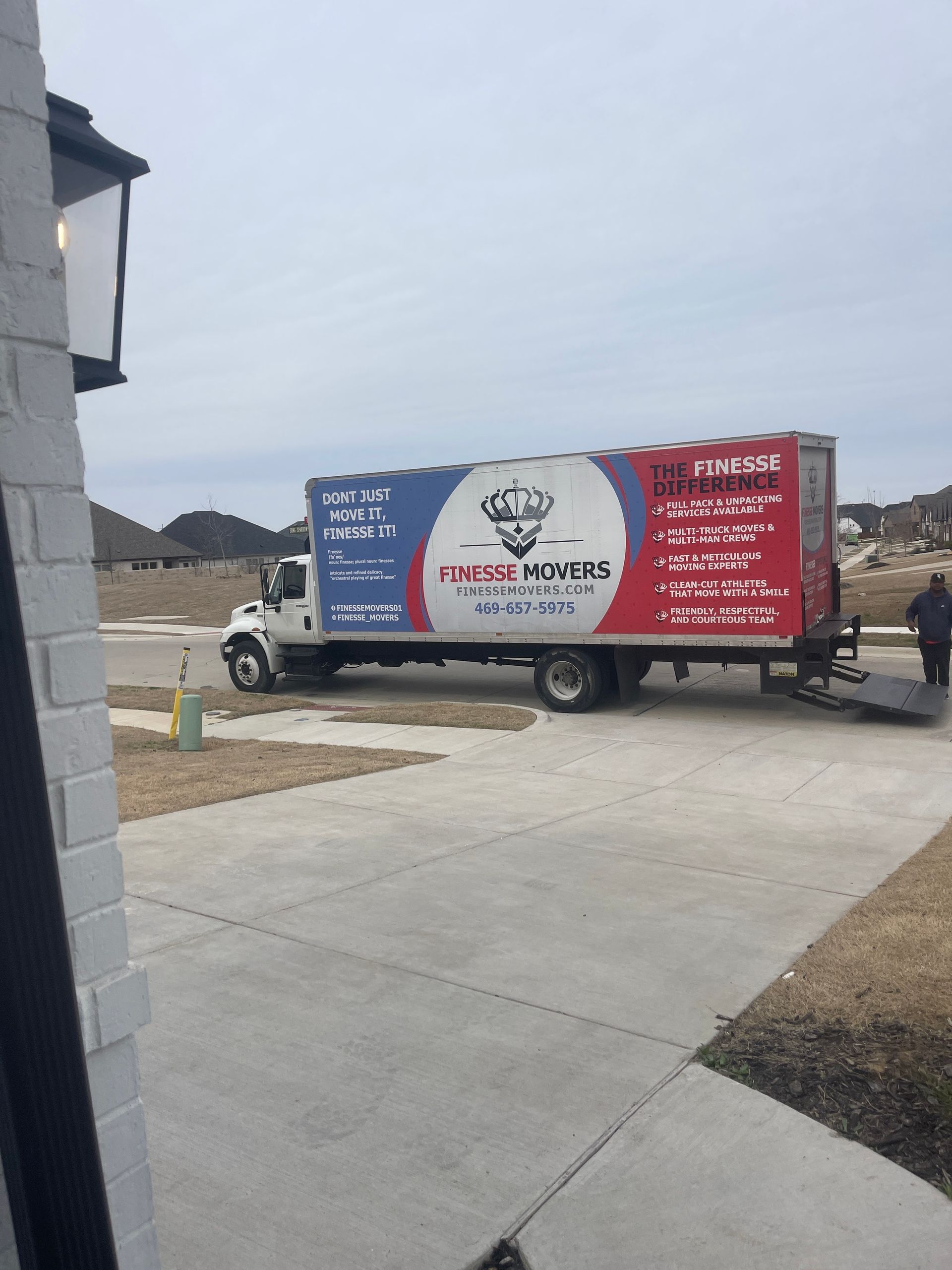 Moving truck parked in a driveway; a person stands near the ramp. The truck has the