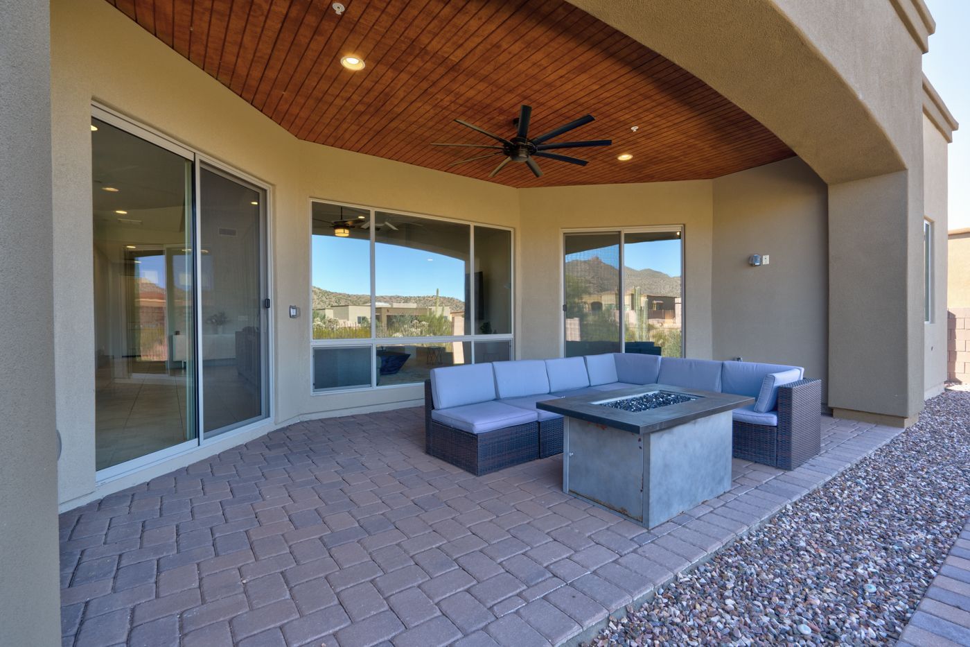 Patio with blue sectional, square fire pit, and glass sliding doors. Beige walls, wood ceiling, and brick pavers.