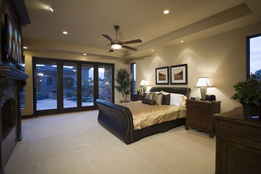 Luxurious bedroom with king-sized bed, fireplace, and sliding doors leading to a patio. Warm lighting and neutral colors.