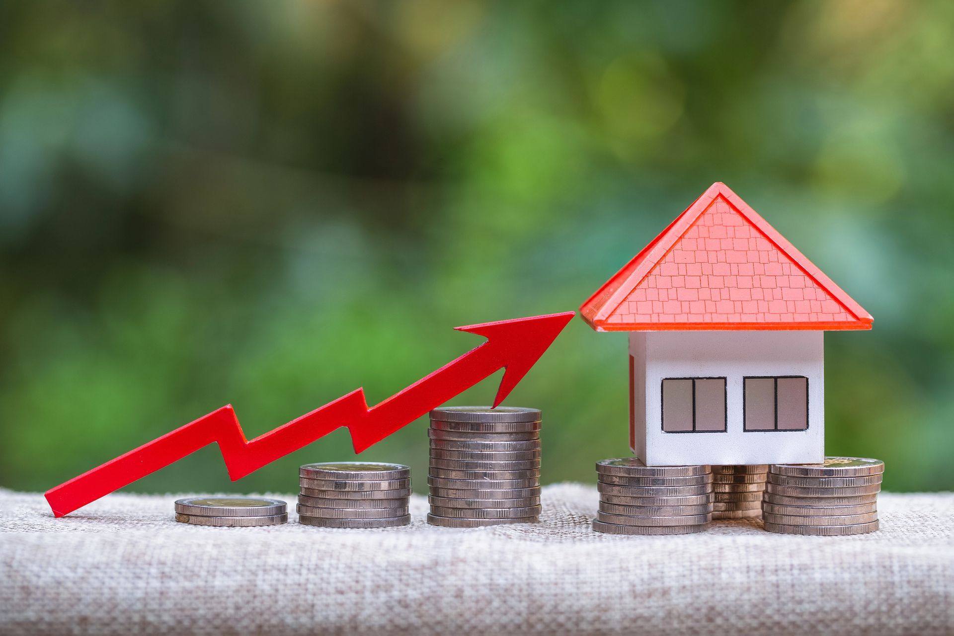 Toy house with red roof on coin stacks, a red arrow pointing upwards, symbolizing rising real estate prices.
