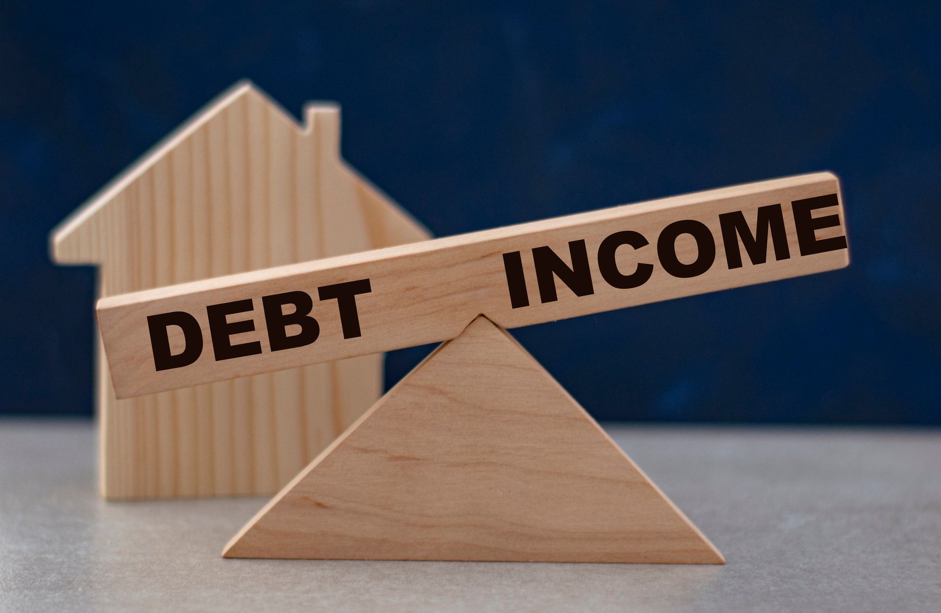 Wooden seesaw balancing DEBT and INCOME; house shape in the background.